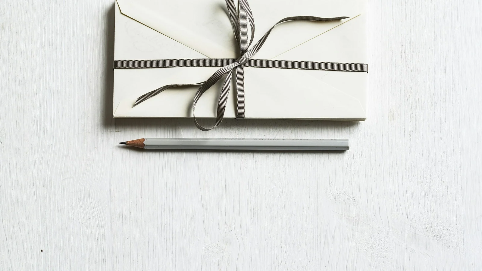 A white envelope, pencil and ribbon arranged on a bright neutral surface.